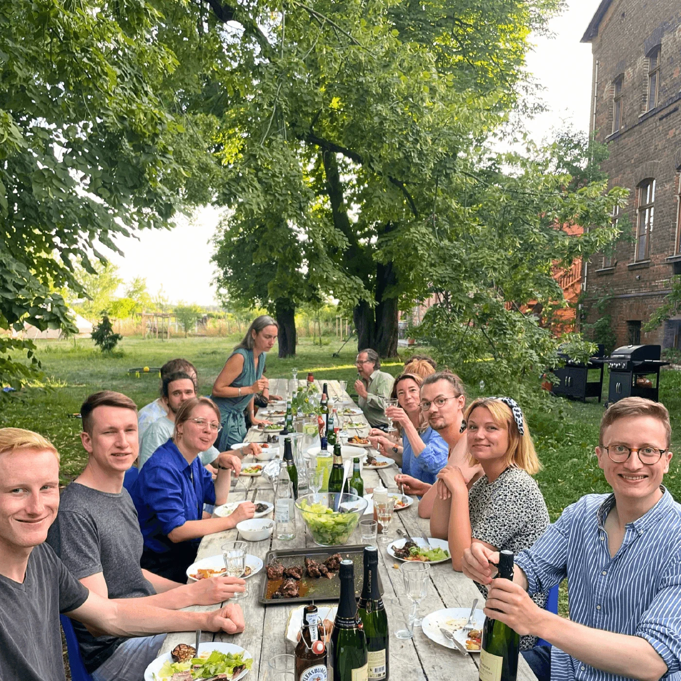 Das Team an einer langen Tafel unter alten Bäumen neben einem Backsteinhaus
